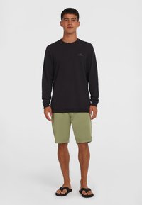 Camicia nera a maniche lunghe con logo sul petto, abbinata a pantaloni corti verde chiaro e infradito neri, in piedi su uno sfondo neutro.