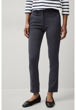 Marineblauwe tailored broek met een slim fit, voorzien van een tailleband met lussen, voorzakken en een cropped lengte. Zwarte platte schoenen. Gestreept topje zichtbaar.