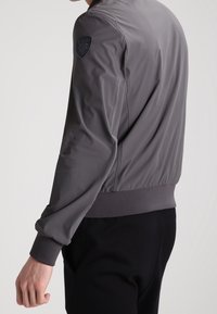 Gray bomber jacket with a smooth, lightweight texture, ribbed cuffs and waistband, and a logo patch on the left shoulder.