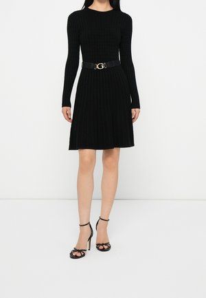 Frau trägt ein schwarzes, langärmeliges Strickkleid mit Taillengürtel und schwarzen High-Heel-Sandalen, steht vor einem schlichten weißen Hintergrund.