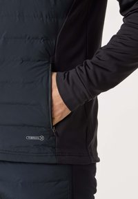 Persoon die een zwarte Thermal-X jas draagt met de rechterhand in een met rits afgesloten zijzak, tegen een neutrale achtergrond.