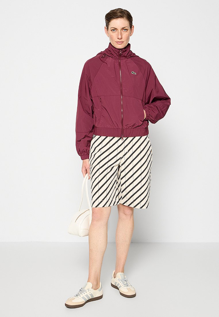 Bordeaux windbreaker met een ritssluiting aan de voorkant, elastische manchetten en een verstelbare capuchon, gecombineerd met een gestreepte crème en zwarte rok tot de knie en sneakers.