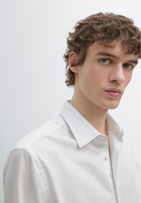 Wit katoenen overhemd met een gestructureerde kraag, knopen aan de voorkant en een gladde textuur, gedragen door een model met krullend haar tegen een lichte achtergrond.