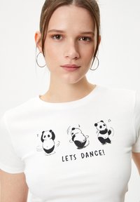 Camiseta blanca ajustada con textura de canalé, que presenta tres ilustraciones de pandas en blanco y negro y el texto "¡BAILEMOS!" debajo.