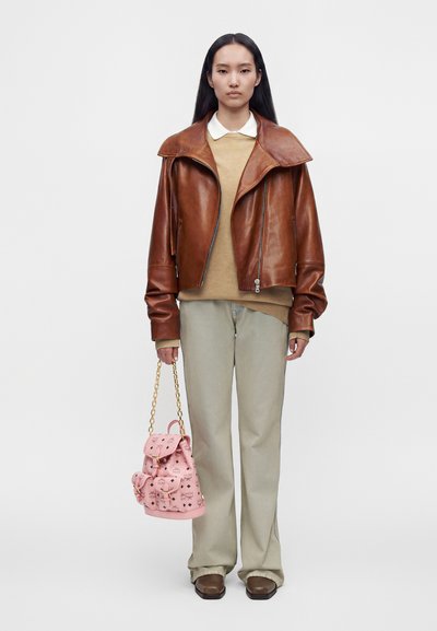 Femme portant une veste en cuir marron, un pull beige, un pantalon clair et des chaussures marron, tenant un sac à main rose à motifs avec une anse chaîne dorée.