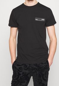 Svart bomullströja med korta ärmar, med en rektangulär logotypklistermärke på övre vänstra sidan. Slät textur och klassisk passform.