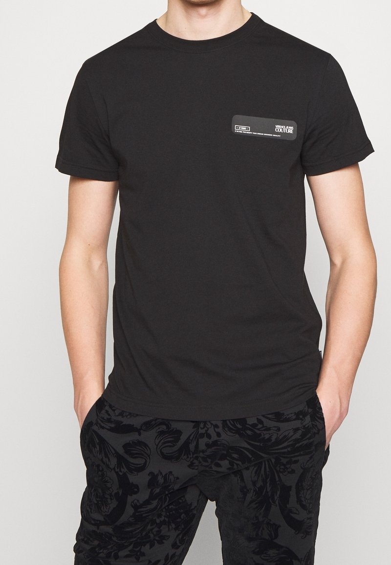 Svart bomullströja med korta ärmar, med en rektangulär logotypklistermärke på övre vänstra sidan. Slät textur och klassisk passform.