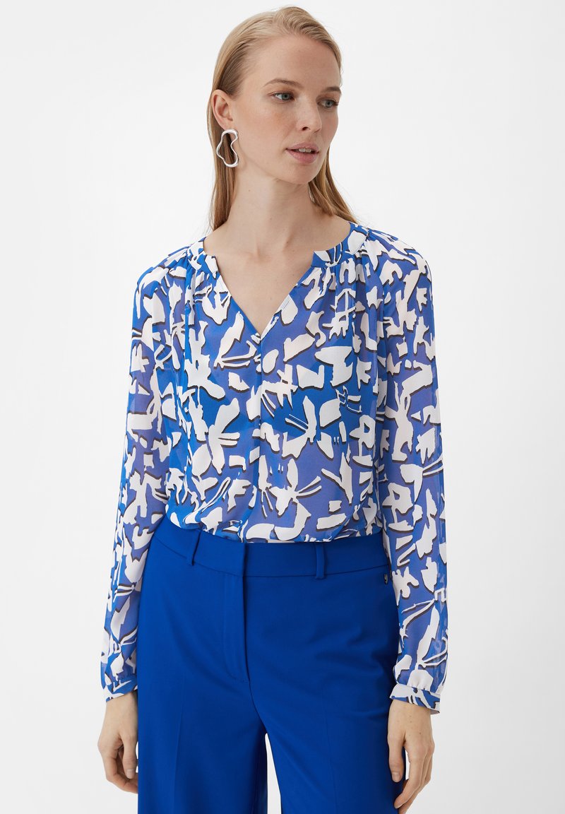 Blaue Bluse mit Muster, V-Ausschnitt, langen Ärmeln und weißen abstrakten Designs. Kombiniert mit maßgeschneiderten blauen Hosen für einen stimmigen Look.