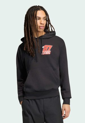 Młoda osoba z długimi dredami w czarnej bluzie z kapturem, na której na piersi znajduje się czerwono-biały napis „#87” oraz „LKNRG”, ręce w kieszeniach.