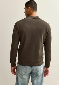 Maglione marrone con una texture liscia, polsini a coste e un orlo dritto. Indossato sopra jeansazzurri chiari, visto da dietro.