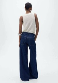 Žena s krátkými vlasy svázanými do drdolu, oblečená v bezrukávovém béžovém pleteném topu, širokých tmavě modrých džínách, stříbrných kruhových náušnicích a náramcích, zobrazená zezadu.