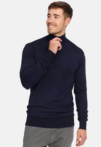 Pull en maille bleu marine avec un col montant, texture côtelée et détail de fermeture éclair. Ajusté avec des manches longues et une taille serrée.
