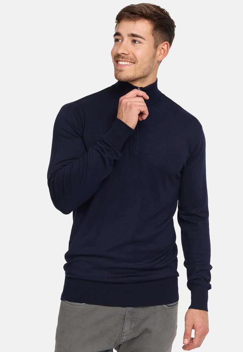 Pull en maille bleu marine avec un col montant, texture côtelée et détail de fermeture éclair. Ajusté avec des manches longues et une taille serrée.
