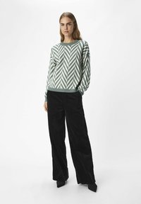 Suéter a rayas en verde claro y blanco con cuello redondo, combinado con pantalones de pata de elefante de pana negra y zapatos de punta negra.