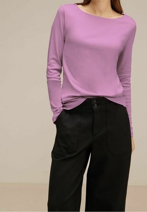 Kobieta ubrana w lawendowy top z długim rękawem, wsunięty w czarne, szerokie spodnie z wysokim stanem, z przednimi kieszeniami i guzikami.