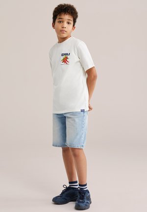 Poiss seisab käed selja taga, seljas valge tšilli graafilise pildiga t-särk, hele teksapüksid, triibulised sokid ja mereväesinised tennised.