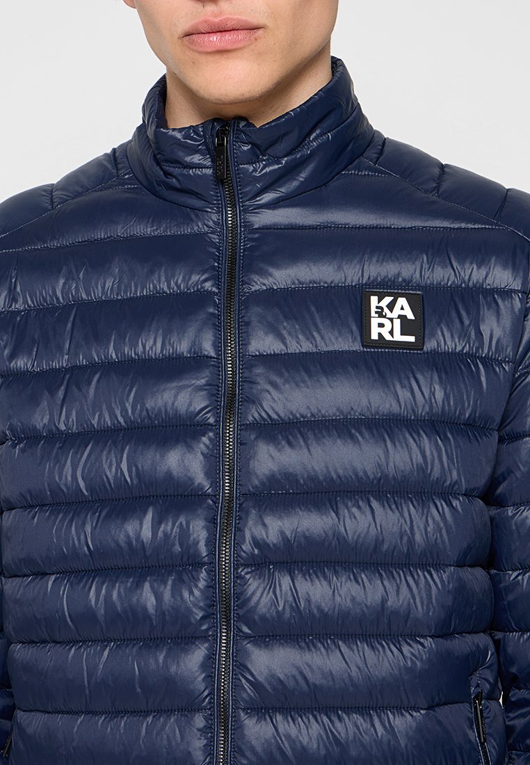 Hombre con una chaqueta acolchada azul marino con cremallera frontal y parche con el logo "KARL" en el pecho.