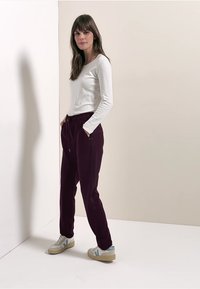 Bílý top s dlouhým rukávem v kombinaci s bordo tepláky s bočními kapsami. Modelka má na sobě světlé tenisky s modrými detaily.