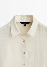 Linen button-up shirt in off-white with a classic collar, six pearl buttons, and a smooth, lightweight texture. Tag visible inside collar.