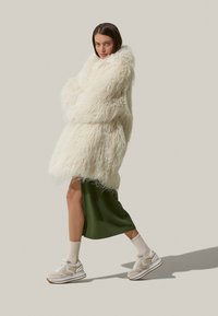 Cappotto in pelliccia sintetica bianco oversize con maniche lunghe, abbinato a un vestito sottoveste verde in satin e sneaker beige. Leggero e soffice.