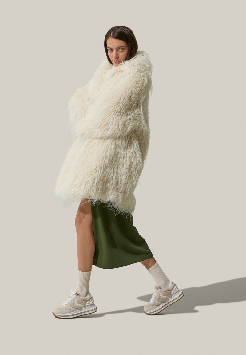 Cappotto in pelliccia sintetica bianco oversize con maniche lunghe, abbinato a un vestito sottoveste verde in satin e sneaker beige. Leggero e soffice.