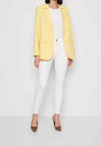 Żółta dopasowana marynarka, biała dopasowana bluzka, białe wąskie dżinsy i brązowe buty na obcasie w wężowy nadruk. Marynarka ma dwie przednie kieszenie i luźny krój.