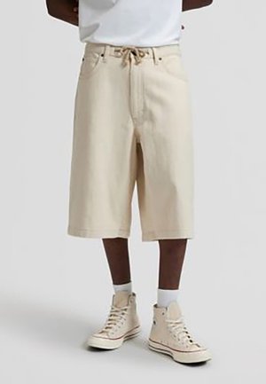 Beige knielange Shorts aus robustem Stoff, mit Kordelzugbund, Vorderentaschen und lockerem Schnitt. Getragen mit hohen Sneakers.