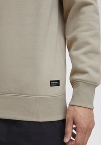 Sudadera beige con puños y dobladillo acanalados. Presenta una etiqueta negra en la parte inferior izquierda. La tela parece suave con un acabado texturizado.