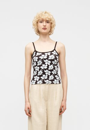 Blond person iført svart og hvitt blomstrete spaghetti-stropptopp og beige vidbukser, stående mot en ensfarget lys bakgrunn.