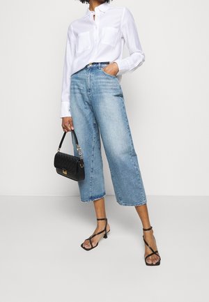 Žena v bílé košili s knoflíky, širokých zkrácených modrých džínách, černých sandálech na podpatku, držící černou kabelku na jednotném pozadí.