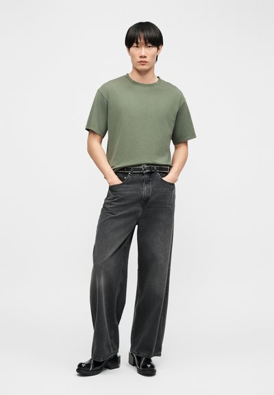 Mladý muž stojaci s rukami v kapsách, oblečený v olivovozelenej tričku, čiernych širokých džínsach s opaskom so cvokmi a čiernych platformových topánkach.