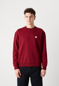 Felpa in cotone bordeaux con colletto e polsini a costine, caratterizzata da una piccola etichetta con logo sul petto. Abbinata a pantaloni scuri. Design semplice.