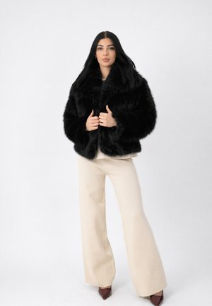 Frau mit langen dunklen Haaren, die eine schwarze Kunstpelzjacke, cremefarbene Hosen mit weitem Bein und braune spitze Schuhe trägt, steht vor einem schlichten Hintergrund.