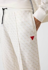 Weiße Jogginghosen aus weichem Stoff mit einem dezenten beigen Muster. Sie verfügen über einen schwarzen Kordelzug und ein rotes dreieckiges Logo auf der Tasche.