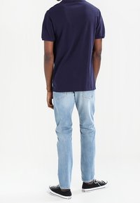 Polo shirt bleu marine à manches courtes, accompagné d'un jean bleu clair et de baskets noires à semelles blanches. Prise de vue par l'arrière.