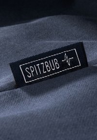 Tessuto blu scuro con una superficie texture, caratterizzato da un'etichetta nera cucita con la scritta "SPITZBUB" in bianco, affiancata da un grafico di una linea del battito. 