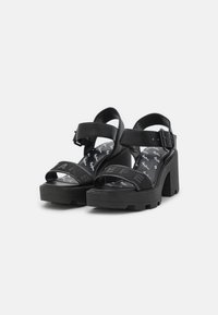 Paire de sandales noires à talons hauts épais avec des brides larges, une bride de cheville avec boucle et une semelle intérieure de marque sur fond blanc.