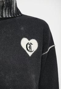Sweater cinza-escuro com um gola alta canelada. Apresenta uma forma de coração branco com a letra "G" preta dentro. Textura suave com ligeira variação de cor.