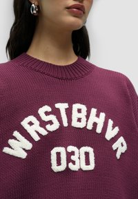 Bordeaux gebreide trui met geribbelde halslijn en witte, textuurvolle letters "WRSTBHLVR 030" die prominent op de voorkant zijn weergegeven.