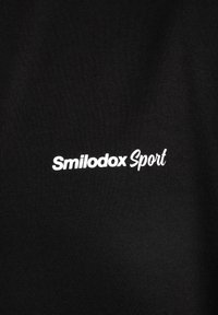 Sort stof med en glat tekstur, der har teksten "Smilodox Sport" i hvid, placeret centreret på beklædningen.