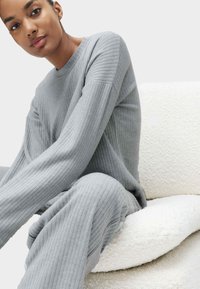 Hellgrauer gerippter Pullover und passende Hose aus weichem Material, kombiniert mit einem strukturierten weißen Stuhl mit einer geschwungenen Form.
