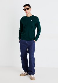 Donkergroen overhemd met lange mouwen met een klein logo op de borst, gecombineerd met losse blauwe spijkerbroeken en tan sneakers. Vierkante zonnebril maakt de look compleet.