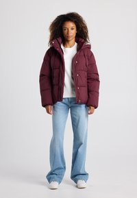 Bordeaux pufferjack met een capuchon, gequilt ontwerp, voorkant met drukknoopsluitingen, gecombineerd met lichtblauwe flared jeans en witte sneakers.