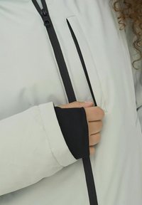 Šviesiai pilkas švarkas su juoda užtrauktuku ir rankovėmis. Tekstūruotas audinys rodo arti kišenės ir rankovės dizaino, sudaryto iš sluoksniuotų medžiagų.