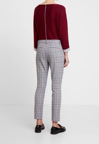 Personne portant un haut bordeaux avec une couture au dos, un pantalon ajusté à carreaux gris, et des mocassins noirs, se tenant contre un fond blanc uni.