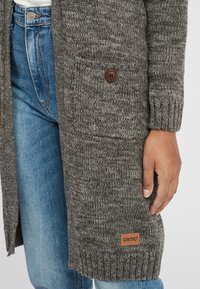 Long, knitted cardigan in dark gray, featuring a button closure, ribbed cuffs, a pocket, and a brown logo patch on the side.