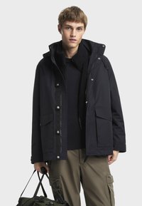 Crna vanjska jakna s kapuljačom, zatvaranjem na patentni zatvarač, dva prednja džepa i unutrašnjim podsuknjama od umjetnog krzna. Model drži zelenu torbu; bež vojne hlače upotpunjuju izgled.