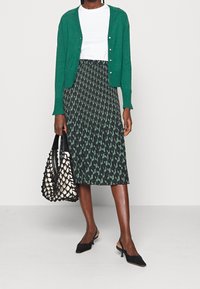 Zelený kardigan přes bílé tričko, doplněný sukní s černým a zeleným geometrickým vzorem. Černé lodičky na podpatku a tkanaá taška dokončují outfit.