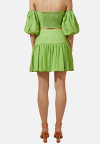 Vestido de dos piezas texturizado en verde, con un top off-shoulder y falda de capas. Acentuado con mangas abullonadas y combinado con sandalias de tacón naranjas.