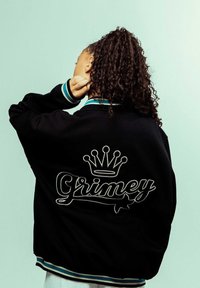 Chaqueta bomber negra con cuello a rayas en color verde azulado, que presenta texto bordado en blanco "Grimey" y un diseño de corona en la parte trasera. Textura suave.